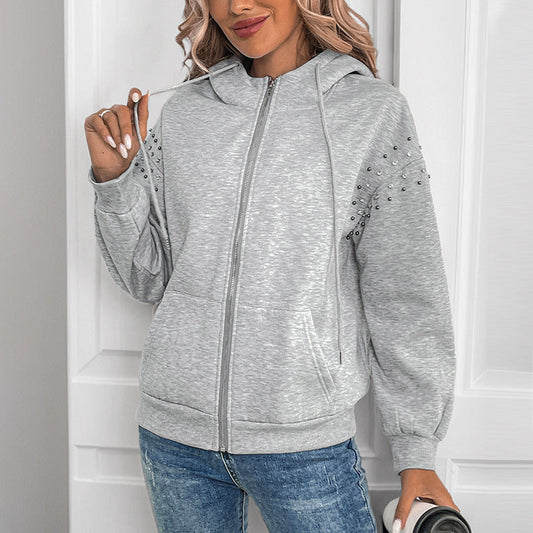 Donna con felpa grigia con cappuccio e borchie, jeans blu, capelli ondulati biondi, sfondo bianco. Moda casual, abbigliamento autunnale.