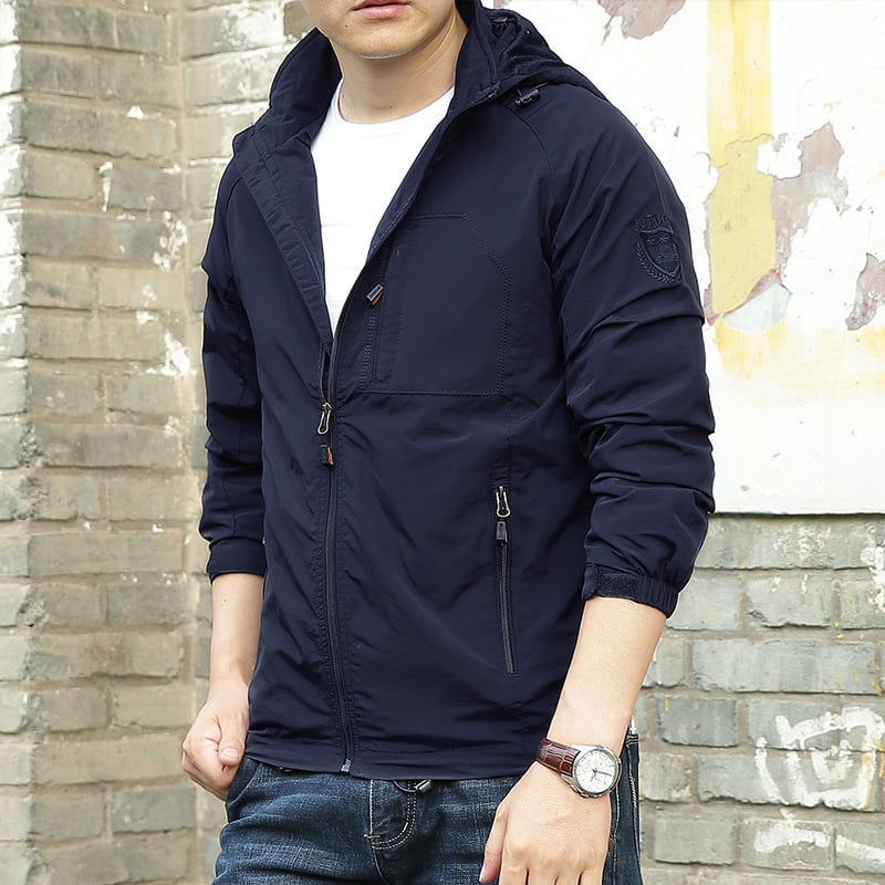 Giacca a vento blu scuro da uomo, con cappuccio e cerniere, indossata su maglietta bianca e jeans. Stile casual, moda maschile autunnale.
