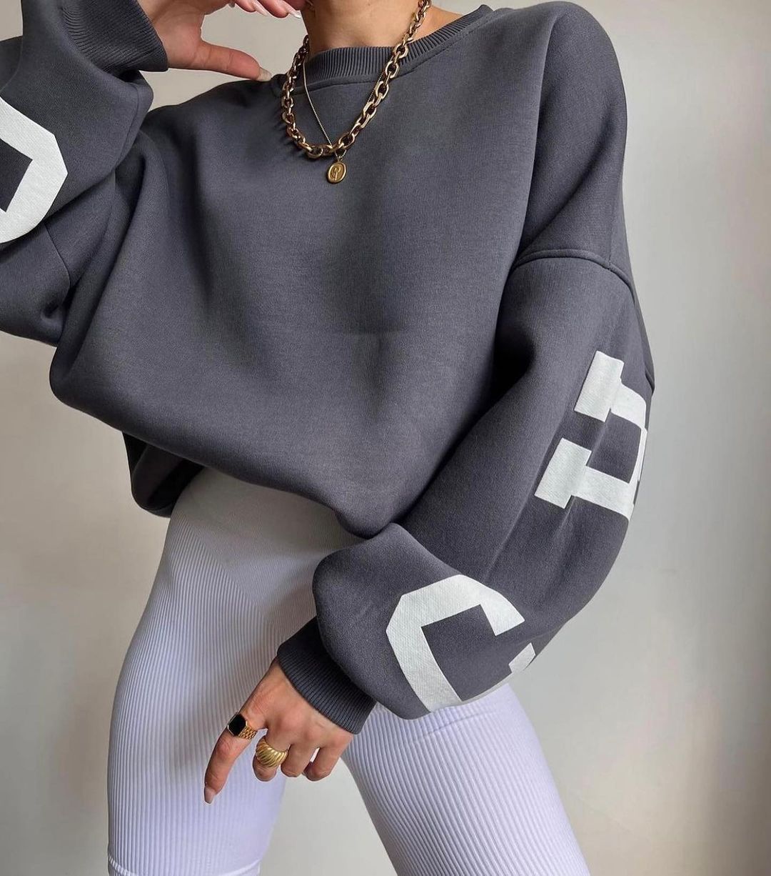 Donna con felpa oversize grigia, dettagli bianchi sulle maniche, collana a catena dorata, leggings bianchi a coste. Moda casual autunnale.