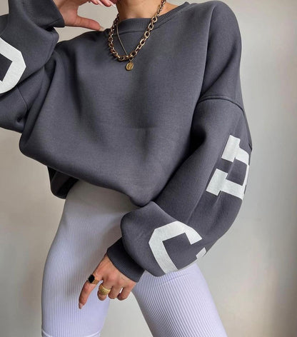 Donna con felpa oversize grigia, dettagli bianchi sulle maniche, collana a catena dorata, leggings bianchi a coste. Moda casual autunnale.