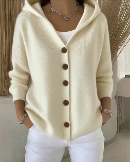 Donna con cardigan bianco con cappuccio e bottoni marroni, indossa pantaloni bianchi. Moda autunnale, abbigliamento casual elegante.