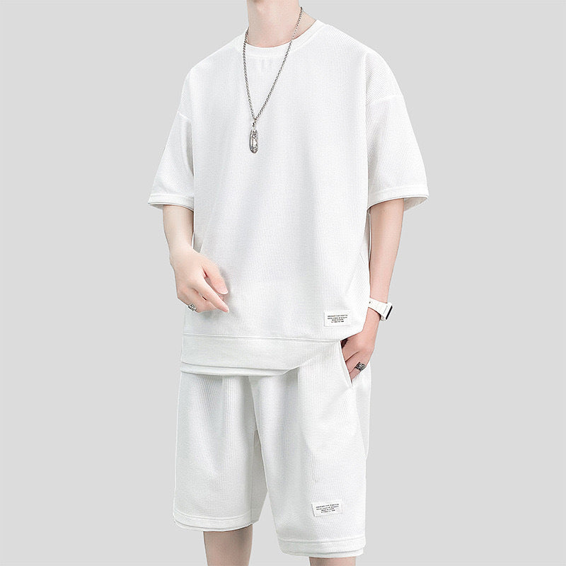 Abbigliamento uomo estivo: completo bianco casual con maglietta oversize e pantaloncini, collana argento, sfondo grigio. Moda streetwear.