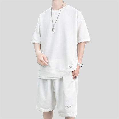 Abbigliamento uomo estivo: completo bianco casual con maglietta oversize e pantaloncini, collana argento, sfondo grigio. Moda streetwear.