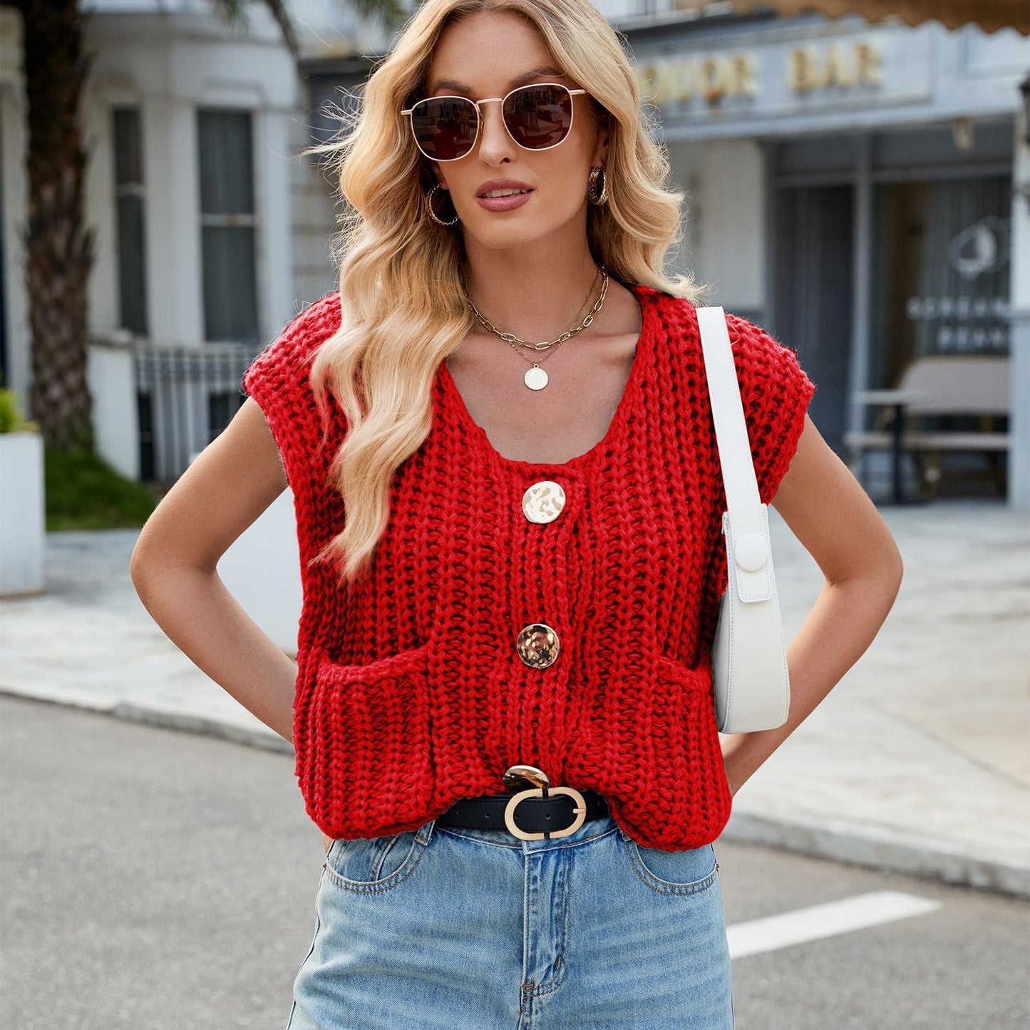 Donna con occhiali da sole indossa maglione rosso a maglia grossa, jeans chiari e borsa bianca. Moda casual, look estivo urbano.