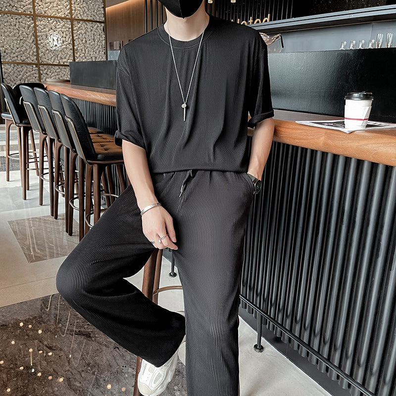 Uomo in abbigliamento casual nero, seduto al bar con caffè. Indossa collana e bracciali, scarpe bianche. Stile moderno ed elegante.