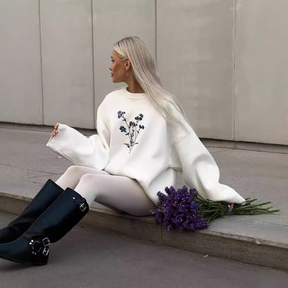Donna con maglione bianco decorato con fiori blu, seduta su marciapiede. Indossa stivali neri alti e tiene un mazzo di fiori viola. Moda autunnale.