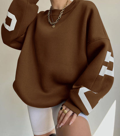 Donna con felpa marrone oversize, maniche con numeri bianchi, indossa collana dorata e anelli, pantaloncini ciclisti bianchi. Moda casual autunnale.