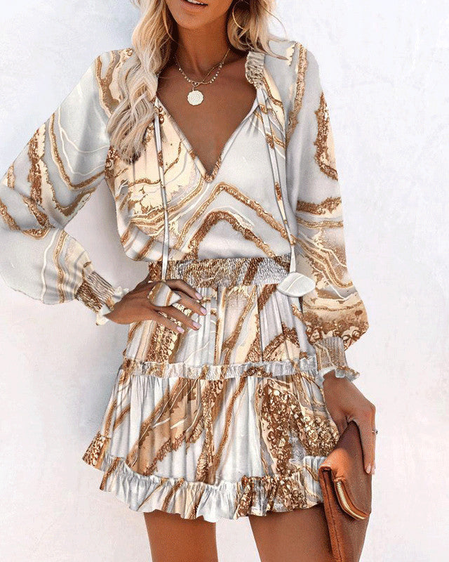 Donna con abito elegante a fantasia marmo beige e bianco, maniche lunghe, gonna a balze, collana dorata, borsa marrone. Moda femminile chic.