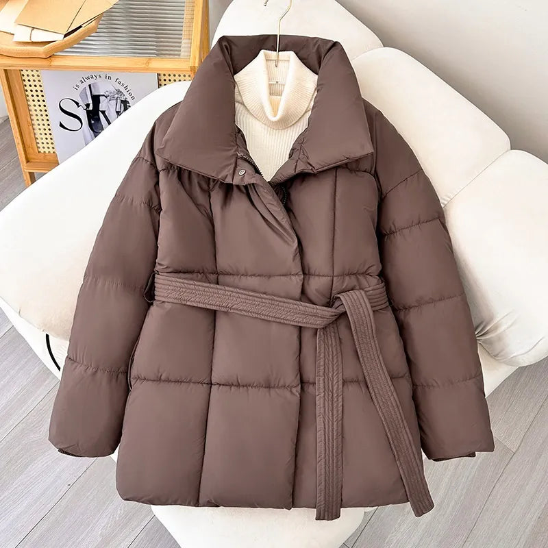 Giacca imbottita marrone con cintura, design trapuntato, su sedia bianca. Moda invernale, abbigliamento caldo, stile casual elegante.