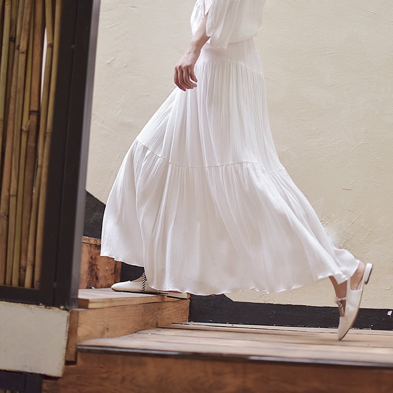 Donna con gonna lunga bianca plissettata scende le scale in legno. Moda estiva elegante, abbigliamento femminile, stile casual chic.