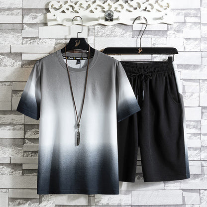 Completo estivo uomo: maglietta sfumata bianco-nero e pantaloncini neri appesi su grucce, sfondo muro mattoni grigi. Moda casual elegante.