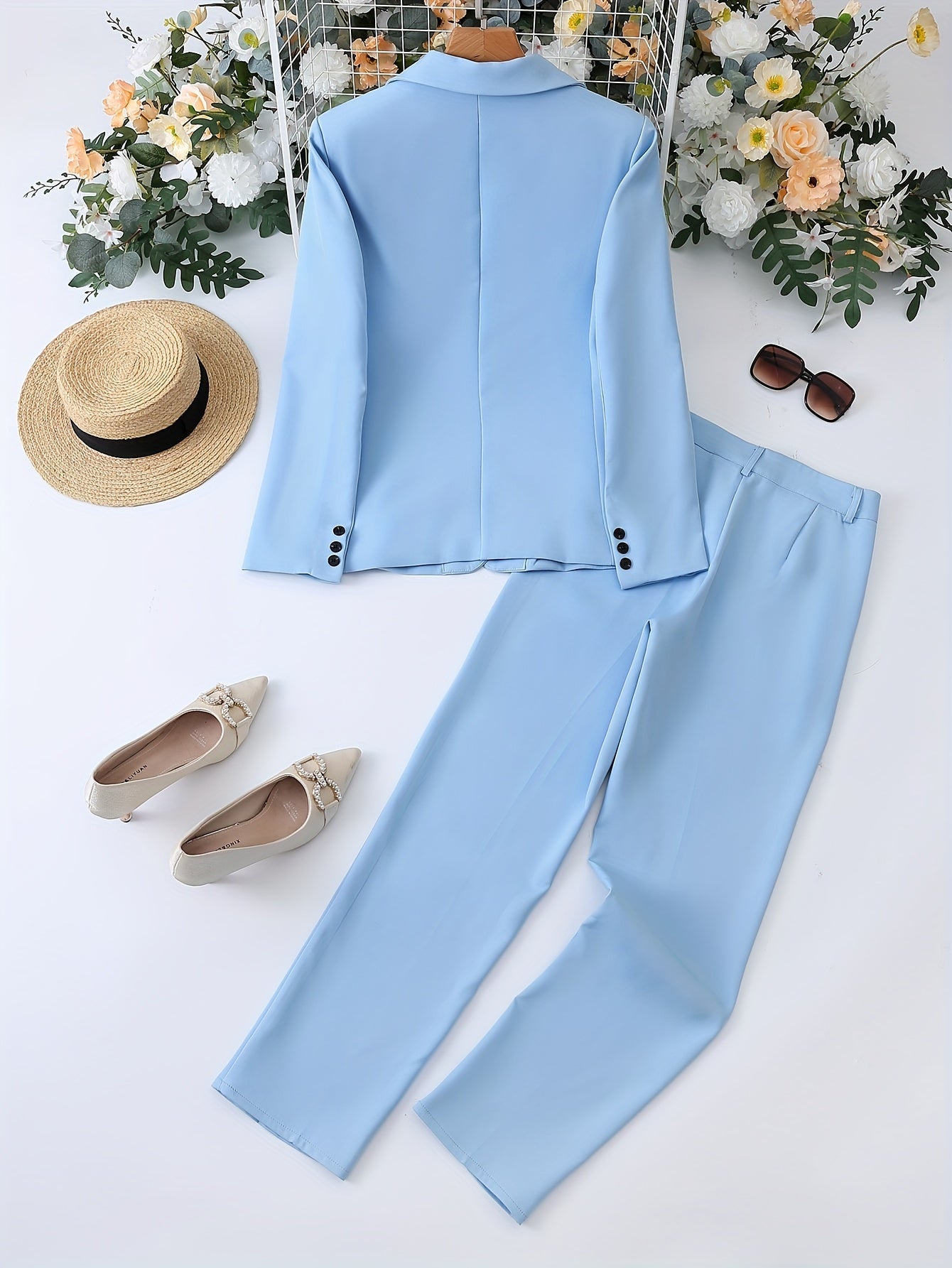 Completo elegante azzurro da donna con giacca e pantaloni, scarpe beige, cappello di paglia e occhiali da sole, su sfondo floreale.