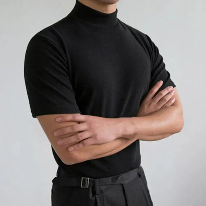 Uomo in maglia nera a collo alto, braccia incrociate, sfondo grigio chiaro. Moda maschile elegante, abbigliamento casual chic.