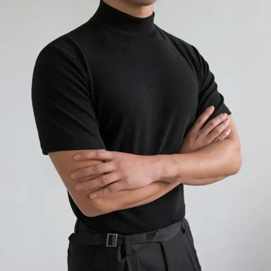 Uomo in maglia nera a collo alto, braccia incrociate, sfondo grigio chiaro. Moda maschile elegante, abbigliamento casual chic.