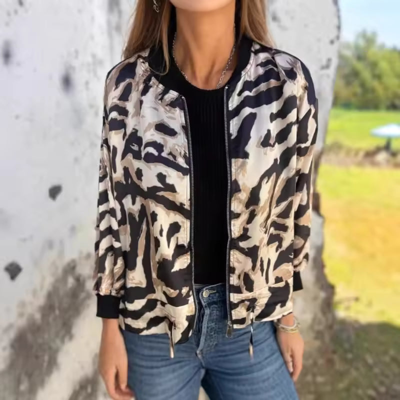 Giacca bomber animalier donna, motivo zebrato nero e beige, abbinata a jeans blu e maglia nera. Moda casual autunnale all'aperto.