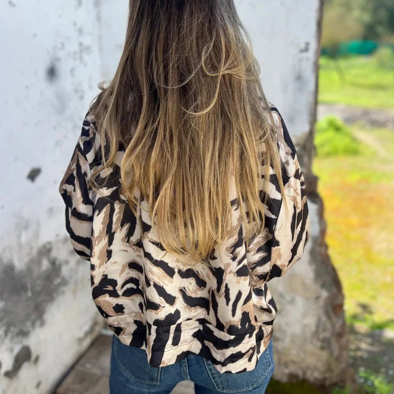 Donna di spalle con capelli lunghi e biondi, indossa camicia a fantasia animalier e jeans. Sfondo esterno con muro bianco e natura verde.