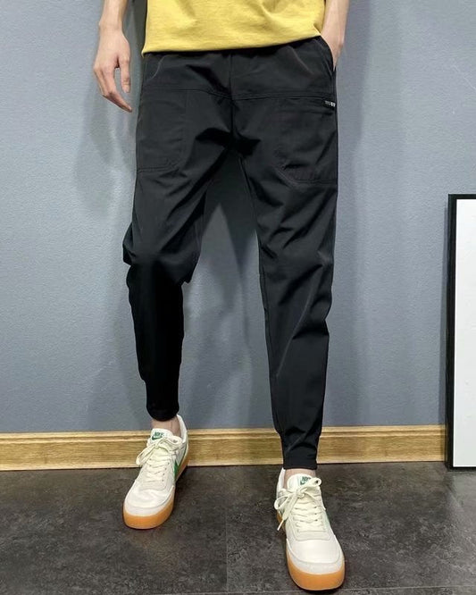 Pantaloni sportivi neri da uomo, abbinati a scarpe da ginnastica bianche con suola marrone, su sfondo grigio. Moda casual maschile.