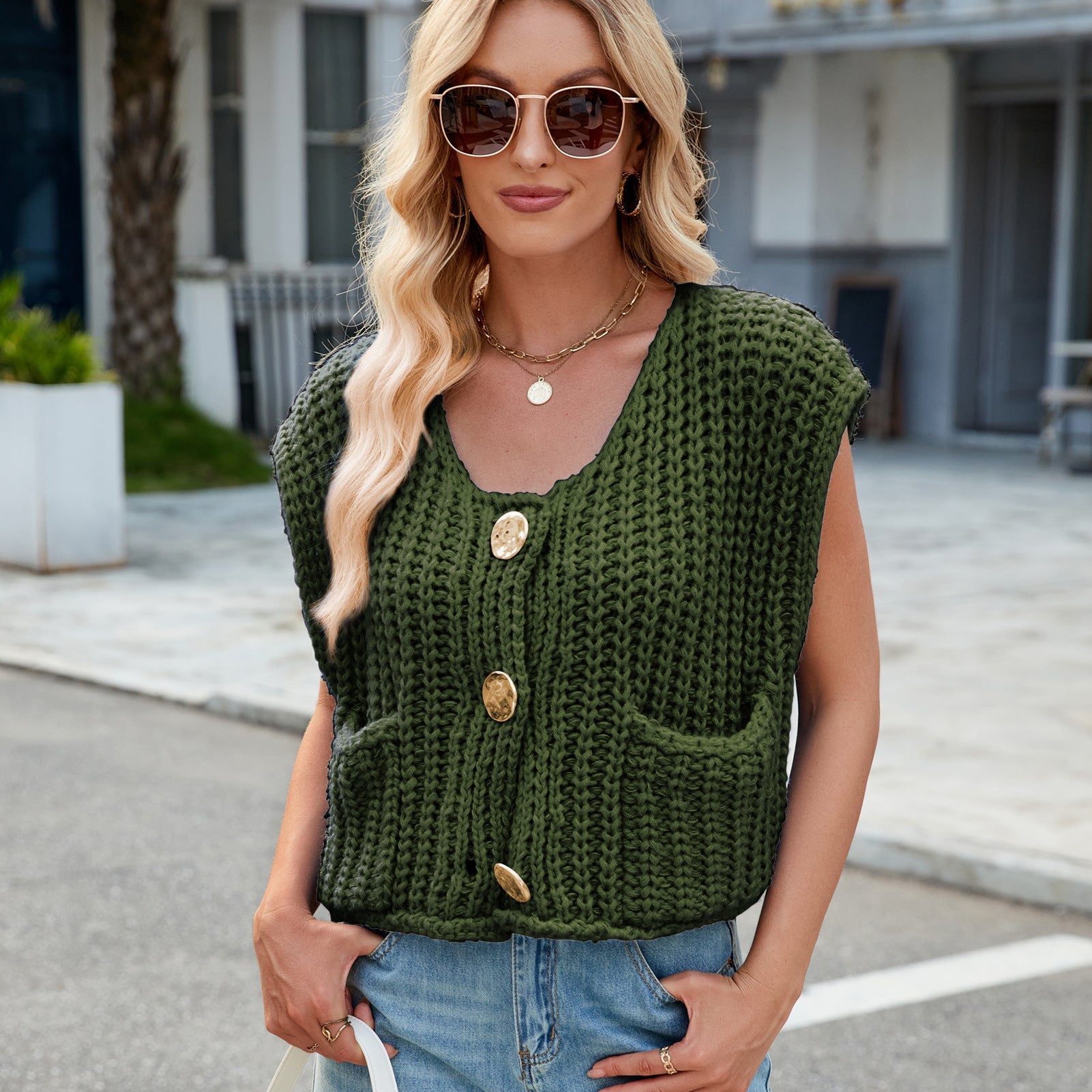 Donna con occhiali da sole indossa un gilet verde a maglia con bottoni grandi e tasche, abbinato a jeans chiari. Moda casual estiva in città.