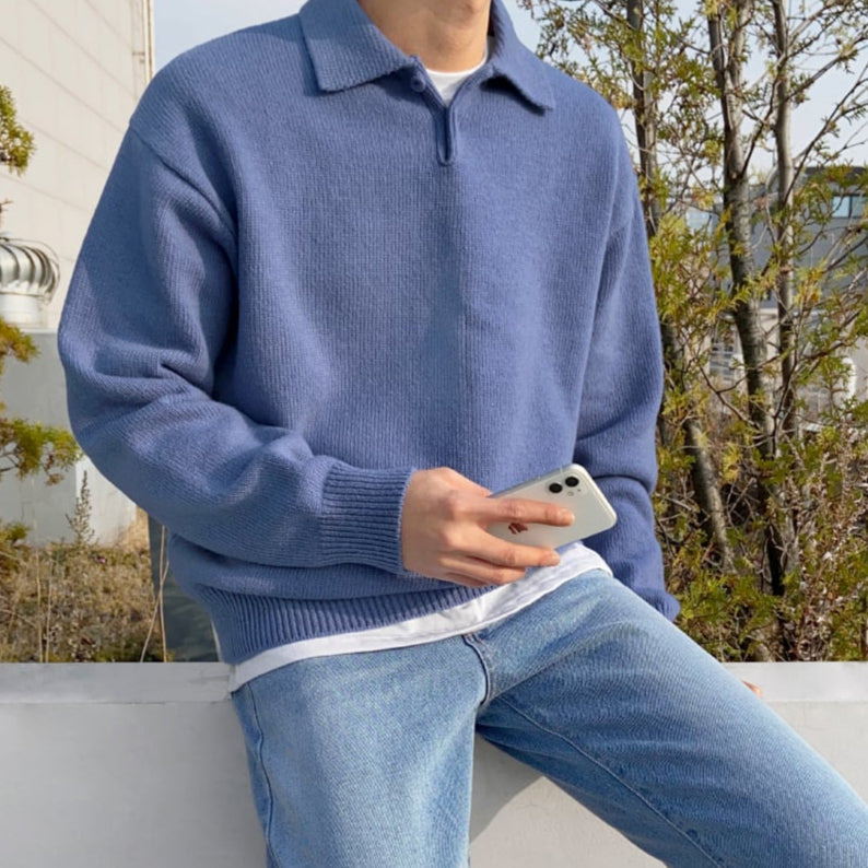 Uomo con maglione blu e jeans, tiene uno smartphone. Sfondo con piante e edificio. Moda casual maschile, tecnologia, abbigliamento autunnale.