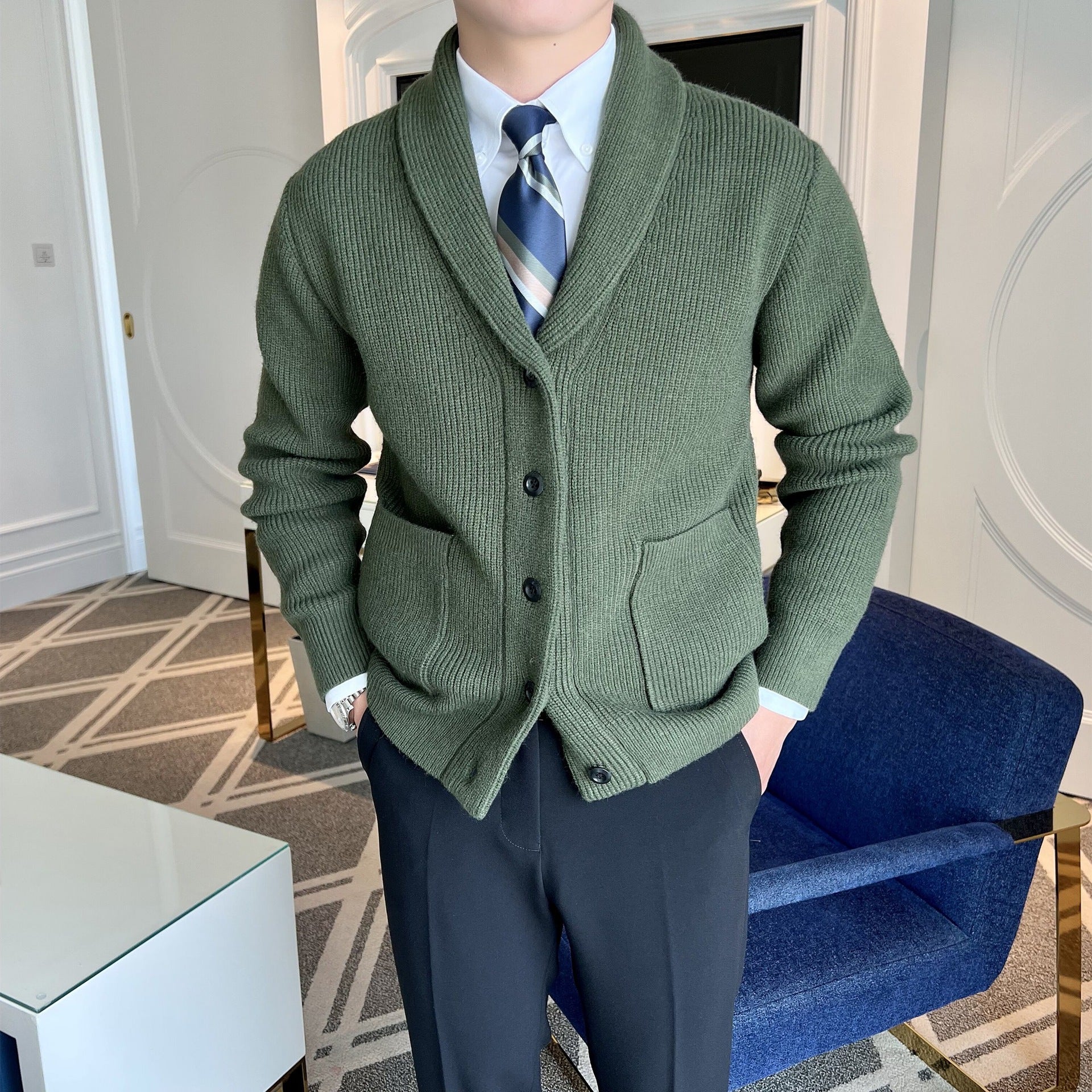 Uomo in giacca di lana verde con cravatta a righe, camicia bianca e pantaloni neri, in un salotto moderno con poltrona blu e tavolino bianco.