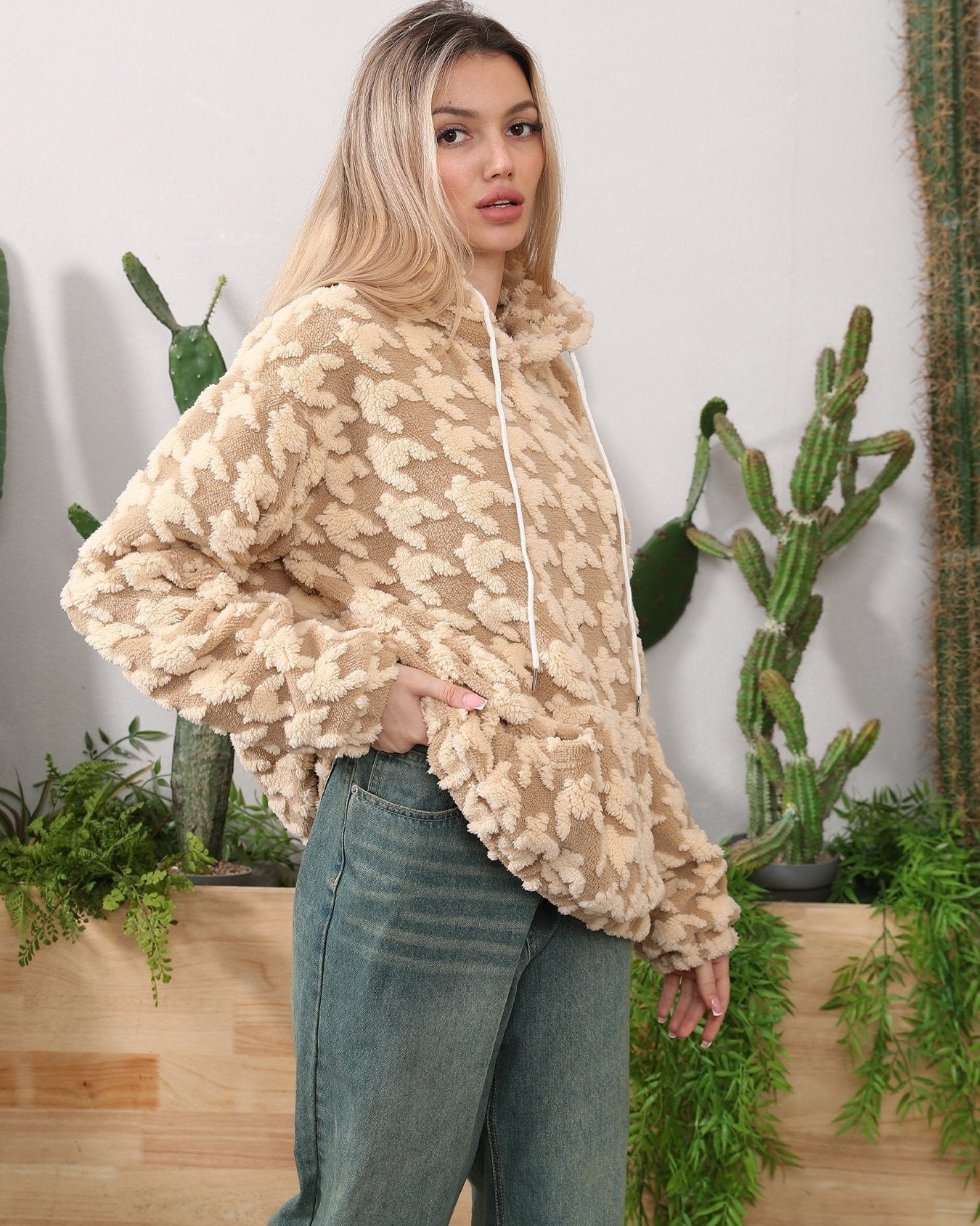 Donna con felpa beige a fantasia pied de poule, jeans, circondata da cactus e piante verdi. Moda casual, abbigliamento autunnale, stile urbano.
