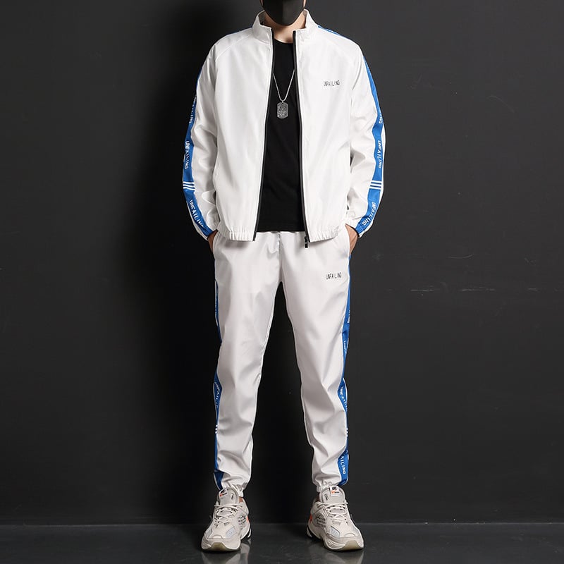 Tuta sportiva bianca con bande blu, indossata da uomo su sfondo nero. Completo casual con scarpe da ginnastica, ideale per moda streetwear.