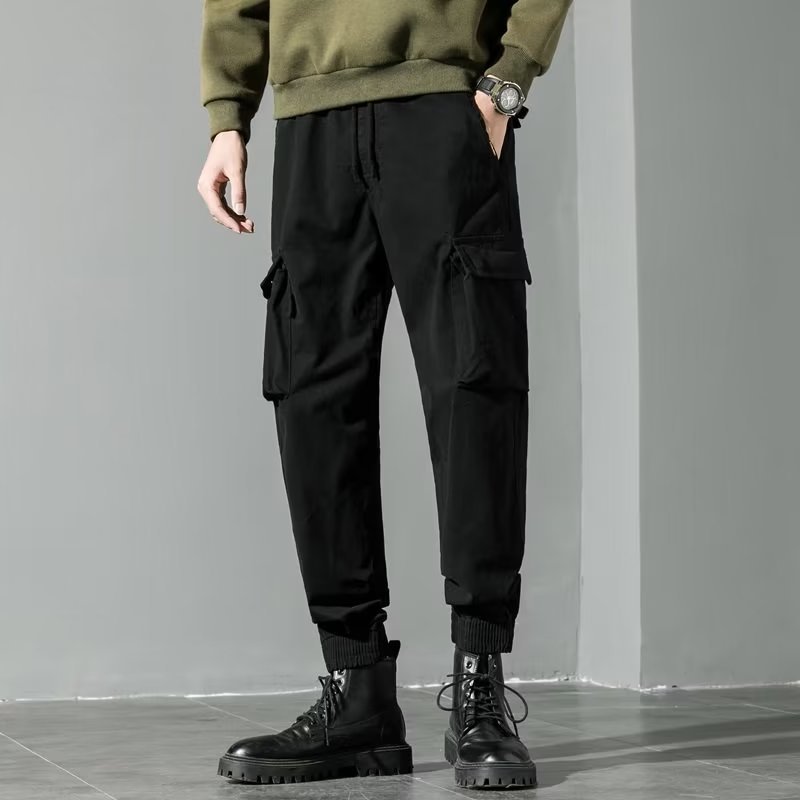 Pantaloni cargo neri da uomo, tasche laterali, abbinati a stivali neri robusti e maglione verde oliva. Moda casual maschile autunno inverno.