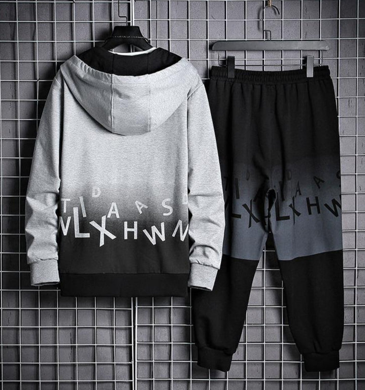 Tuta sportiva uomo grigia e nera con cappuccio, design moderno con lettere, appesa su griglia metallica. Abbigliamento casual alla moda.