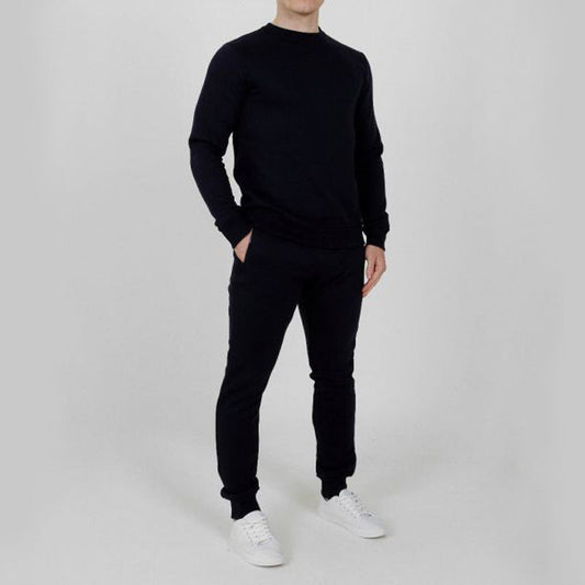Uomo in tuta sportiva nera, con felpa e pantaloni abbinati, indossa scarpe da ginnastica bianche. Sfondo neutro. Moda casual maschile.