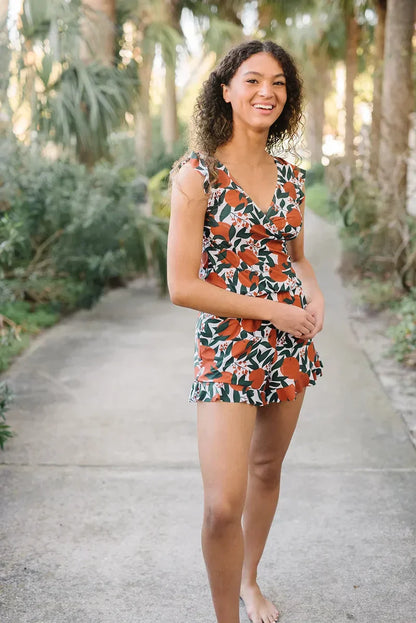 Giovane donna sorridente in abito estivo floreale arancione e verde, cammina a piedi nudi su un sentiero circondato da palme e vegetazione lussureggiante.