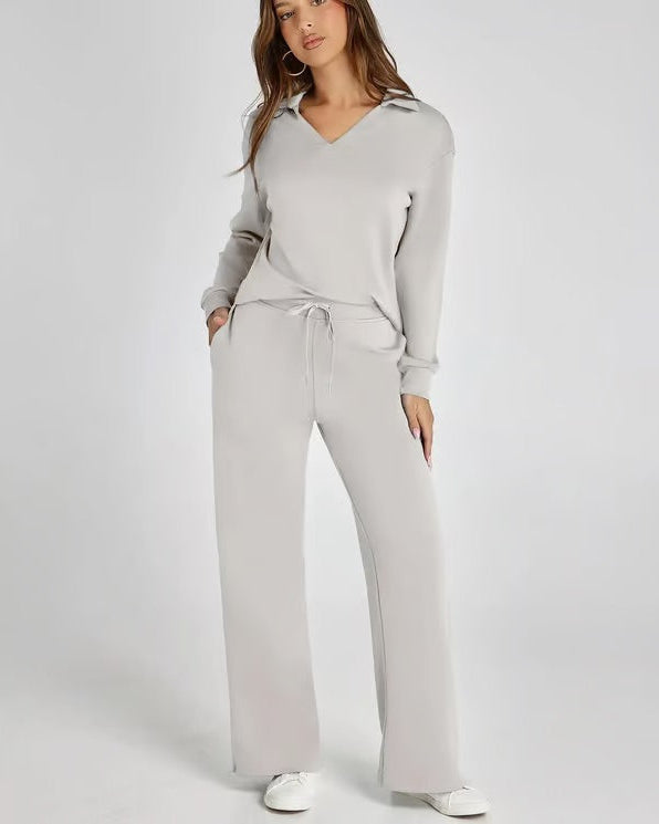Donna in tuta grigia elegante, con maglia a maniche lunghe e pantaloni larghi, posa su sfondo bianco. Abbigliamento casual chic, moda donna.