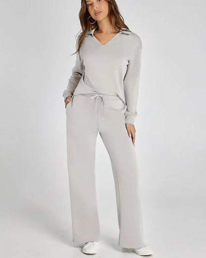 Donna in tuta grigia elegante, con maglia a maniche lunghe e pantaloni larghi, posa su sfondo bianco. Abbigliamento casual chic, moda donna.
