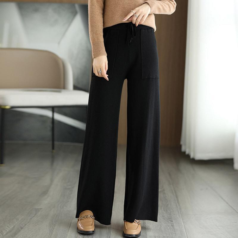 Donna in pantaloni neri larghi, maglione beige, scarpe marroni, in un salotto moderno. Moda autunnale, abbigliamento casual elegante.