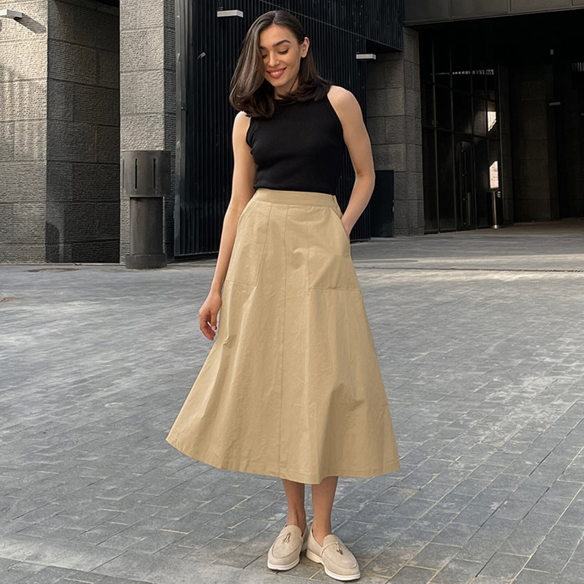 Donna con gonna beige e top nero, posa su strada urbana. Moda casual elegante, scarpe bianche. Sfondo architettonico moderno.