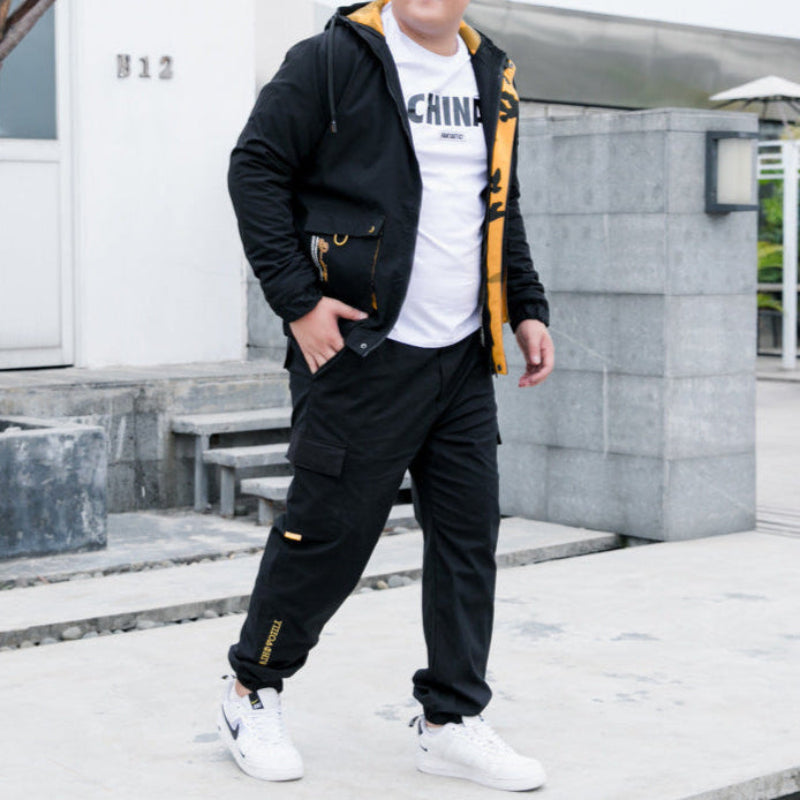 Uomo in tuta sportiva nera con dettagli gialli, giacca aperta su maglietta bianca, scarpe da ginnastica bianche. Moda casual maschile.