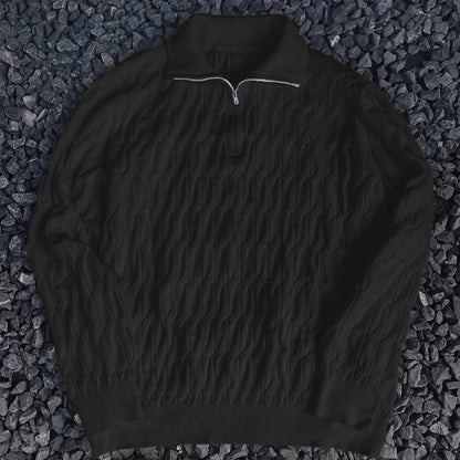 Maglione nero a collo alto con zip, motivo a trecce, su sfondo di pietre scure. Elegante e versatile, ideale per moda autunno-inverno.