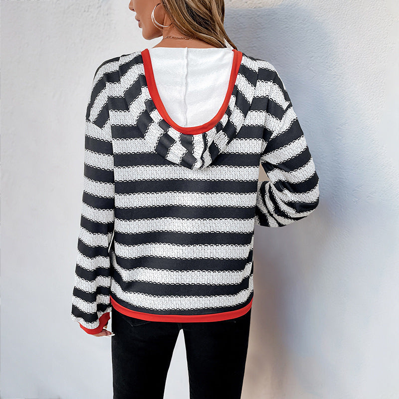Donna di spalle con maglia a righe bianche e nere, cappuccio, bordi rossi, orecchini a cerchio, sfondo bianco. Moda casual autunnale.
