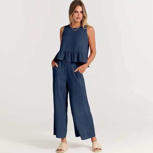 Donna in tuta blu senza maniche con volant, pantaloni larghi, mani in tasca, indossa sandali beige. Moda estiva elegante e casual.