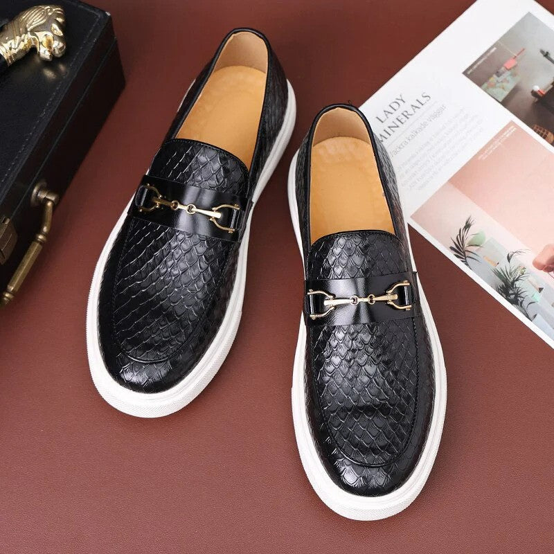Scarpe mocassini in pelle nera con motivo a squame, suola bianca e dettaglio metallico dorato, su sfondo marrone con rivista e pianta decorativa.