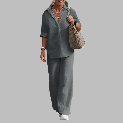 Donna in abbigliamento casual grigio, camicia e pantaloni larghi, con borsa intrecciata beige e scarpe bianche, su sfondo grigio.