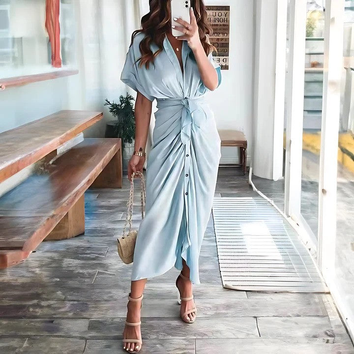 Donna in abito lungo azzurro con cintura, sandali beige e borsa a tracolla, in un ambiente moderno con panchina in legno e pavimento in piastrelle.