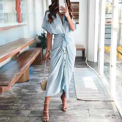 Donna in abito lungo azzurro con cintura, sandali beige e borsa a tracolla, in un ambiente moderno con panchina in legno e pavimento in piastrelle.