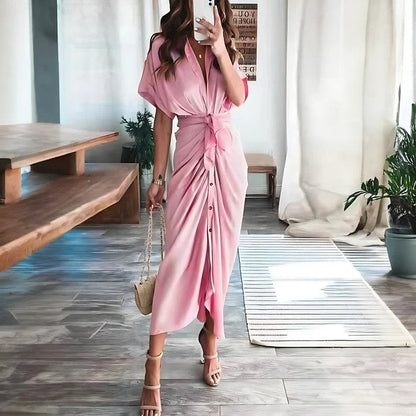 Donna in abito lungo rosa con bottoni, sandali beige e borsa a tracolla, in un salotto moderno con piante e tavolo in legno. Moda estiva elegante.