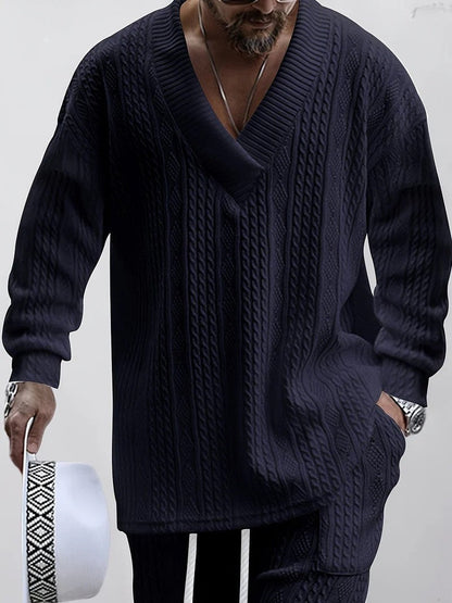 Uomo con maglione a trecce blu scuro, cappello bianco in mano, occhiali da sole, orologio argento. Moda maschile elegante e casual.