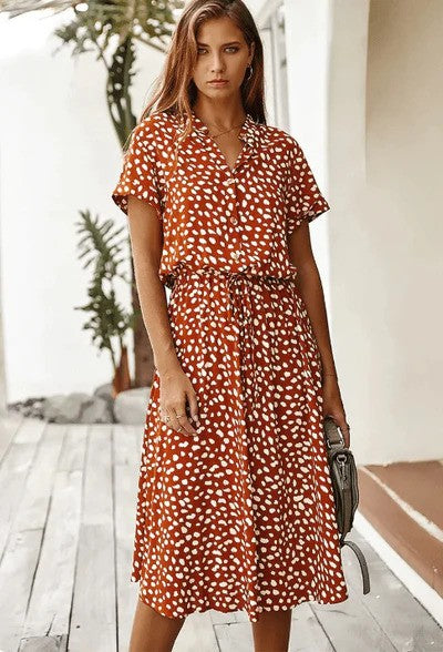 Donna con abito midi a pois marrone e bianco, maniche corte, in piedi su una veranda. Moda estiva, stile casual chic, abbigliamento femminile trendy.