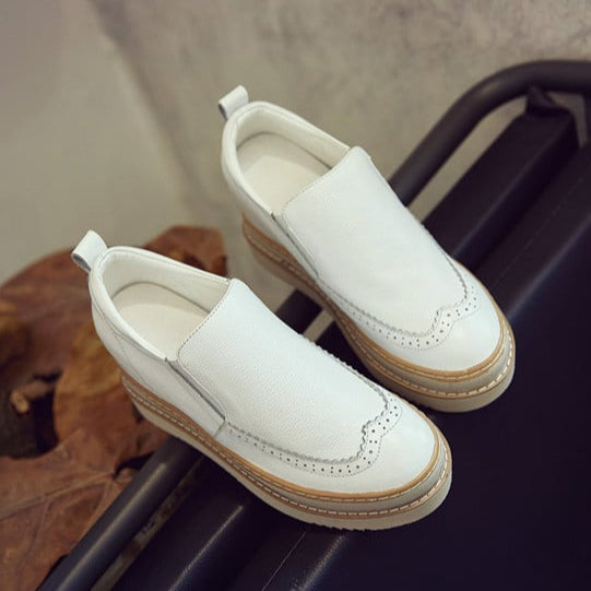 Scarpe slip-on bianche in pelle con suola in gomma marrone, dettagli brogue, su sfondo neutro. Calzature eleganti e comode per ogni occasione.