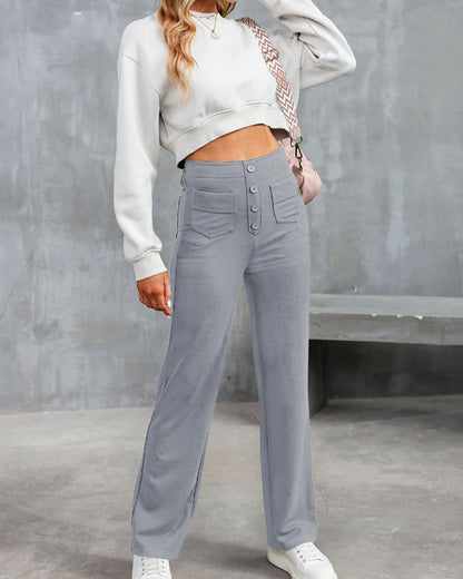 Donna in pantaloni grigi a vita alta con bottoni, maglione bianco corto, borsa a tracolla e sneakers bianche, su sfondo grigio urbano.