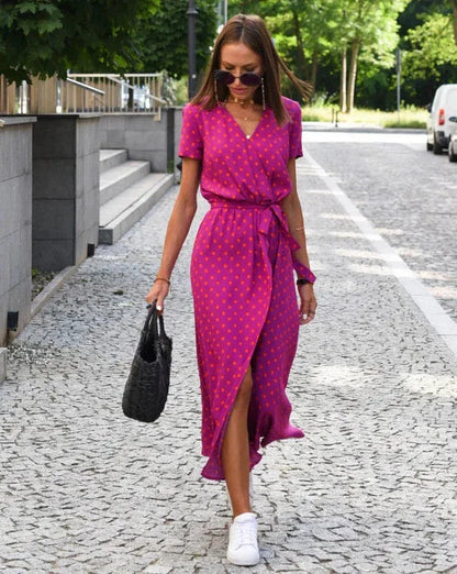 Donna con abito rosa a pois, cammina su strada lastricata, indossa occhiali da sole e scarpe bianche, borsa nera intrecciata, giornata estiva.