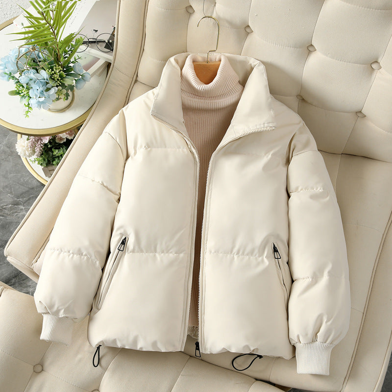 Giacca imbottita bianca su poltrona beige, con zip e tasche, abbinata a maglione a collo alto color crema. Moda invernale elegante e calda.