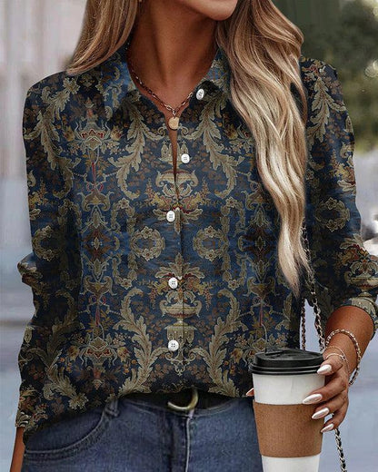 Donna con camicia a fantasia barocca blu e oro, capelli biondi ondulati, tiene una tazza di caffè. Moda autunnale elegante e casual.